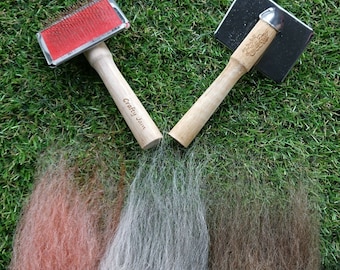 May include: Two wooden pet grooming tools on a green grass background. One is a red and silver brush with the words "Crafty Jane" on the handle. The other is a silver metal comb with a wooden handle.