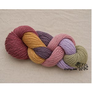 Puede incluir: Una madeja de hilo trenzada con una paleta multicolor de rojo intenso, amarillo dorado, morado oscuro, rosa empolvado, lavanda claro y verde salvia. En la esquina inferior derecha aparece un logotipo gr&aacute;fico negro de una oveja y el texto Crafty Jam.