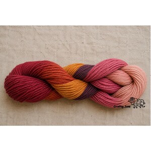 Puede incluir: Una madeja de hilo trenzada con un degradado de colores que incluye rojo intenso, naranja, morado oscuro, magenta y rosa claro. El hilo tiene una textura retorcida e incluye un peque&ntilde;o gr&aacute;fico negro de una oveja con el texto Crafty Jam.