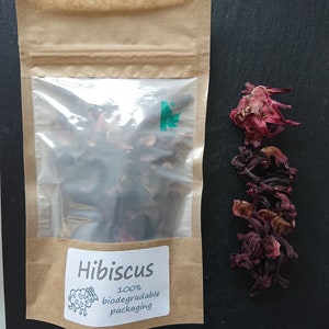 Puede incluir: Una bolsa de papel marr&oacute;n con una ventana de pl&aacute;stico transparente que contiene flores de hibisco secas de color rojo oscuro. La etiqueta dice Hibiscus 100% biodegradable packaging con un dibujo de una oveja. Flores sueltas al lado.