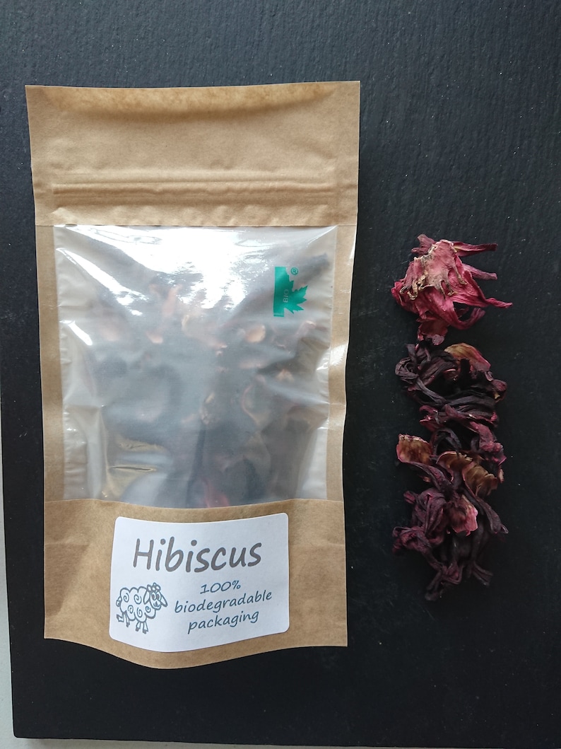 Puede incluir: Una bolsa de papel marr&oacute;n con una ventana de pl&aacute;stico transparente que contiene flores de hibisco secas de color rojo oscuro. La etiqueta dice Hibiscus 100% biodegradable packaging con un dibujo de una oveja. Flores sueltas al lado.