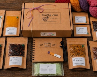 Puede incluir: Una caja de kit de manualidades Crafty Jam atada con cuerda multicolor, rodeada de ocho bolsas de papel marrón que contienen varios tintes naturales y aditivos, un cuaderno de espiral Crafty Notes, una tira reactiva de pH y una madeja de lana multicolor.