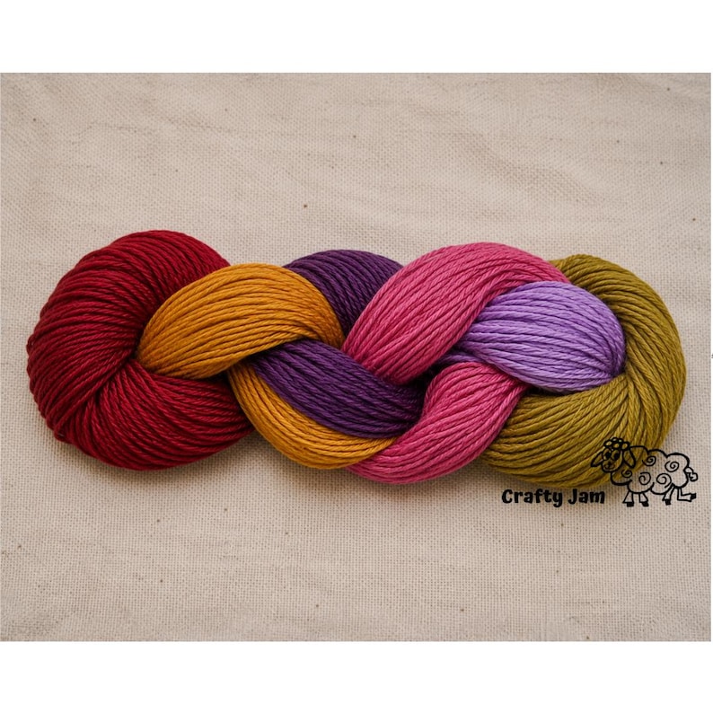 Puede incluir: Una madeja de hilo trenzada con una paleta de colores vibrantes que incluye rojo intenso, amarillo mostaza, morado oscuro, rosa fuerte, lavanda claro y verde oliva. El texto Crafty Jam est&aacute; impreso en negro en la esquina inferior derecha.