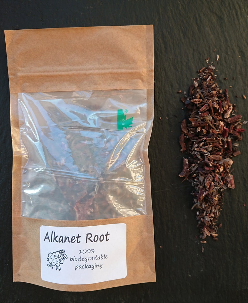 Puede incluir: Una bolsa de papel marr&oacute;n con una ventana de pl&aacute;stico transparente que contiene ra&iacute;z de alcana seca. La etiqueta muestra el texto Alkanet Root, un dibujo de una oveja y el texto 100% biodegradable packaging. Junto a la bolsa hay un mont&oacute;n de trozos de ra&iacute;z le&ntilde;osa de color rojo oscuro.