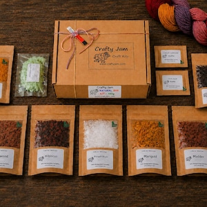 Puede incluir: Una caja de cart&oacute;n marr&oacute;n etiquetada como Crafty Jam Craft Kits con un kit de tinte natural, rodeada de varias bolsas peque&ntilde;as que contienen diversos polvos coloridos y materiales vegetales secos, adem&aacute;s de una madeja de lana multicolor.