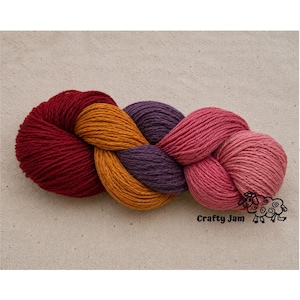 Puede incluir: Una madeja de hilo trenzada que presenta cuatro colores distintos: rojo intenso, amarillo dorado, morado oscuro y rosa. El texto Crafty Jam y un peque&ntilde;o gr&aacute;fico de una oveja est&aacute;n impresos en negro en la esquina inferior derecha.