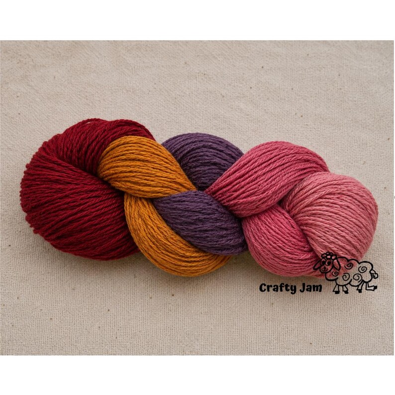 Puede incluir: Una madeja de hilo trenzada que presenta cuatro colores distintos: rojo intenso, amarillo dorado, morado oscuro y rosa. El texto Crafty Jam y un peque&ntilde;o gr&aacute;fico de una oveja est&aacute;n impresos en negro en la esquina inferior derecha.