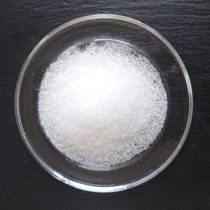 Puede incluir: Un peque&ntilde;o plato redondo de cristal transparente lleno de un mont&oacute;n de gr&aacute;nulos cristalinos blancos y finos. La textura de la sustancia parece granular y reflectante, con part&iacute;culas individuales que captan la luz para crear un brillo intenso.