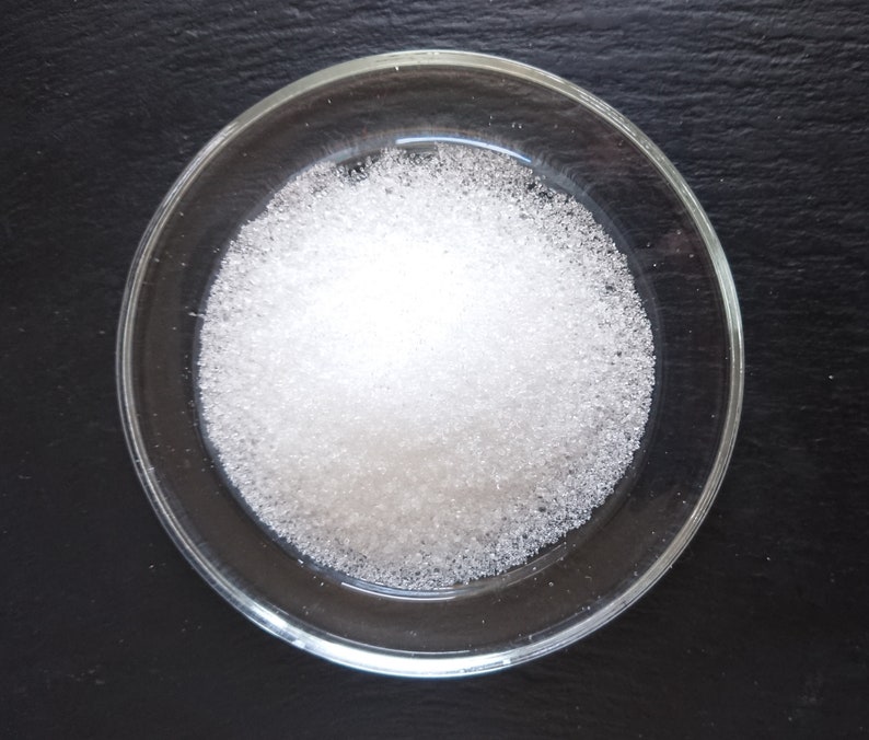 Puede incluir: Un peque&ntilde;o plato redondo de cristal transparente lleno de un mont&oacute;n de gr&aacute;nulos cristalinos blancos y finos. La textura de la sustancia parece granular y reflectante, con part&iacute;culas individuales que captan la luz para crear un brillo intenso.