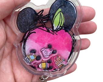 May include: A clear plastic badge holder shaped like Mickey Mouse's head. The holder has a pink and white watercolor design inside, with a black outline of Mickey Mouse's head. There are several small, colorful, cartoon-like images inside the holder, including a school bus, a backpack, a pencil, and a stack of books.