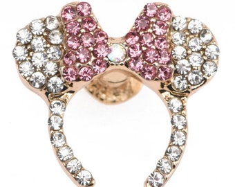 Könnte beinhalten: Ein goldfarbenes Stirnband mit einer Schleife aus rosa und klaren Strasssteinen. Das Stirnband ist mit klaren Strasssteinen verziert.