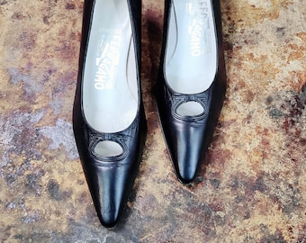 May include: A pair of black leather Salvatore Ferragamo pumps. The shoes have pointed toes and a small heel. The vamp features a cut-out detail with the brand's signature logo. The insoles are white with the brand name printed in gold.