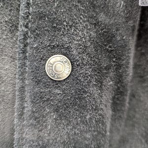 May include: Close-up of a dark grey suede jacket. A silver button with the word "SCHOTT" is visible. The jacket has visible stitching and a textured surface.