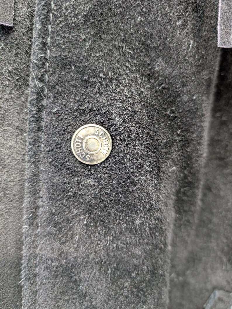 May include: Close-up of a dark grey suede jacket. A silver button with the word "SCHOTT" is visible. The jacket has visible stitching and a textured surface.