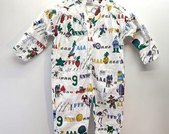 May include: White baby jumpsuit with a colourful all-over print of letters, numbers, and whimsical illustrations. The long-sleeved jumpsuit has a V-neck, button closure, and two front pockets. The garment is hanging on a wooden hanger.