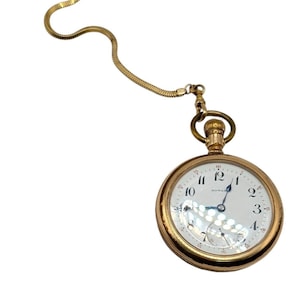 Könnte beinhalten: Eine goldfarbene Taschenuhr mit weißem Zifferblatt, schwarzen Ziffern und blauen Zeigern. Die Uhr hat ein kleines Sekundenrad und ist an einer goldenen Kette befestigt. Ein klassischer Zeitmesser.