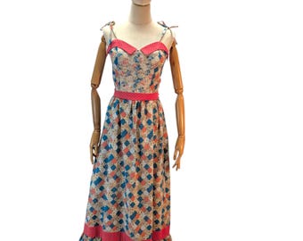 May include: A vintage-style maxi dress with a floral and geometric pattern. The dress features a fitted bodice with a pink trim, tie straps, and a flowing skirt with a ruffled hem. The dress is displayed on a wooden mannequin.