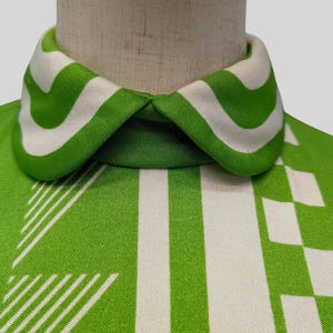 May include: Close-up of a vintage green and white patterned top. The collar features bold green and white stripes. The fabric has a geometric design with vertical stripes and square patterns. The top is displayed on a mannequin.