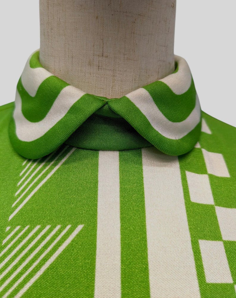 May include: Close-up of a vintage green and white patterned top. The collar features bold green and white stripes. The fabric has a geometric design with vertical stripes and square patterns. The top is displayed on a mannequin.