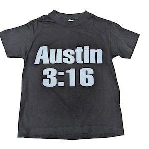 May include: Black t-shirt with the text "Austin 3:16" in light blue block letters. The shirt is short-sleeved and has a crew neck. The text is centered on the front of the shirt.
