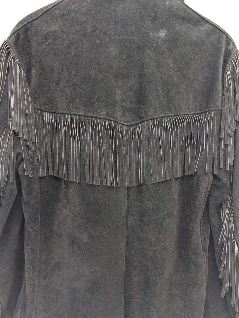 May include: Black suede jacket with fringe detailing across the back and sleeves. The jacket has a classic western style with a notched collar and a button-up front. The fringe is long and hangs down from the yoke and sleeves.