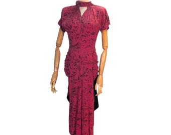 May include: A vintage red dress with a black pattern, displayed on a wooden mannequin. The dress features short sleeves, a keyhole neckline, and a ruched design. The dress is knee-length and has a black accent on the side.