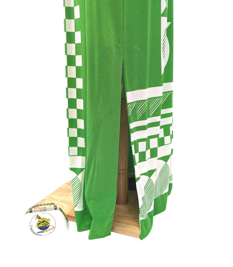 May include: A green garment with white geometric patterns, including checks and angled lines, is displayed. The fabric is draped over a wooden structure, showcasing the design's details. The garment's design is visible, with a split down the center.