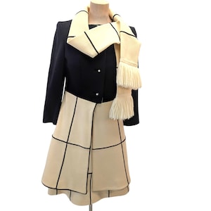 Puede incluir: Un conjunto vintage con una chaqueta negra y una falda color crema. La falda tiene un patrón de cuadrícula delineado en negro. Una bufanda color crema con ribetes negros y flecos está drapeada sobre la chaqueta. El atuendo se exhibe en un maniquí.