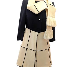 May include: A vintage ensemble featuring a black jacket with a cream-colored skirt. The skirt has a grid pattern outlined in black. A cream-colored scarf with black trim and fringe detail is draped over the jacket. The outfit is displayed on a mannequin.