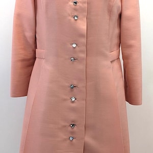 May include: A peach-colored long-sleeved coat featuring a classic collar and a vertical row of ten circular, clear crystal-like buttons down the center front. The garment has a structured silhouette with long sleeves and a smooth, solid fabric finish.