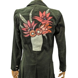 Puede incluir: Una chaqueta de pana verde oscuro con una aplicación floral colorida en la espalda. La aplicación presenta un jarrón con flores en tonos de rosa, naranja y azul.