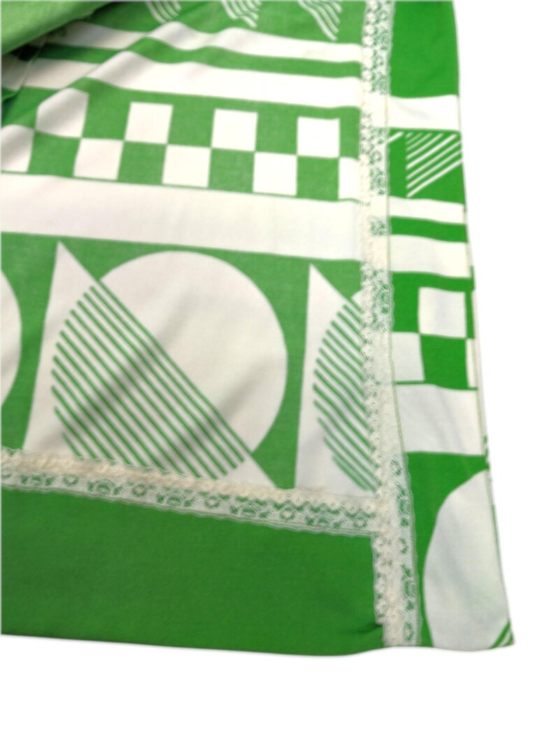 May include: A section of a green and white patterned textile. The design includes geometric shapes like squares, circles with striped segments, and lines. A white lace trim borders the edge of the fabric.