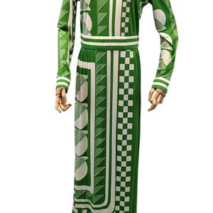May include: Green and white patterned maxi dress. The long-sleeved dress features a collared neckline and a fitted waist. The design includes geometric shapes, including circles, squares, and diagonal lines. The dress is displayed on a wooden mannequin.