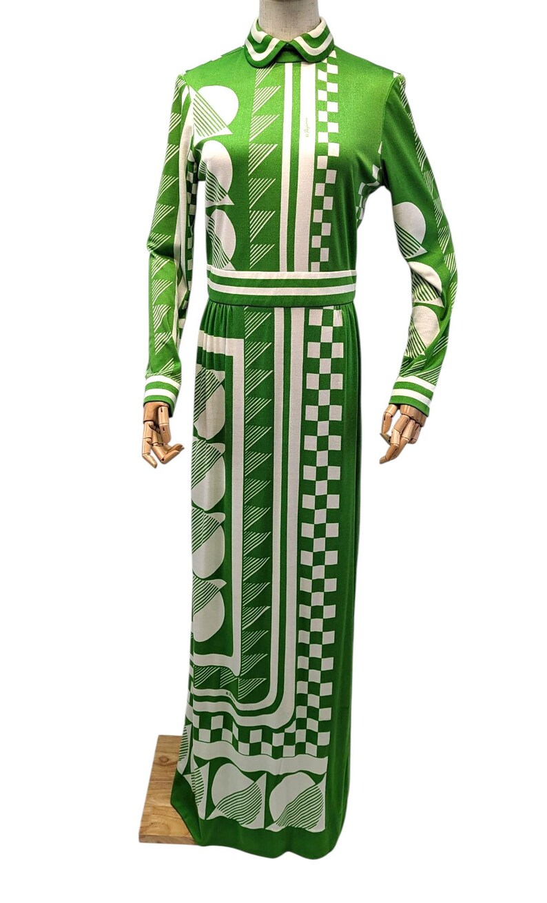 May include: Green and white patterned maxi dress. The long-sleeved dress features a collared neckline and a fitted waist. The design includes geometric shapes, including circles, squares, and diagonal lines. The dress is displayed on a wooden mannequin.