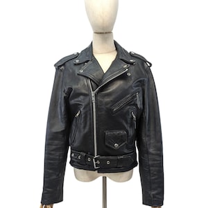 Könnte beinhalten: Schwarze Leder-Motorradjacke mit Reverskragen, asymmetrischem Reißverschluss und mehreren Taschen. Die Jacke hat Schulterklappen, einen Gürtel in der Taille und silberfarbene Hardware. Sie wird auf einer Schaufensterpuppe vor weißem Hintergrund präsentiert.