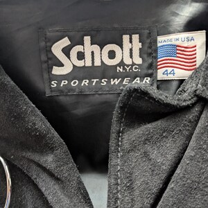 May include: Close-up of a black suede jacket's label. The label reads "Schott N.Y.C. SPORTSWEAR" in white lettering. A small tag displays the American flag and the number "44".