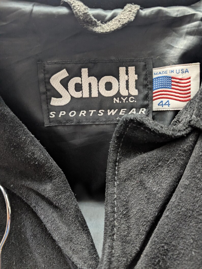 May include: Close-up of a black suede jacket's label. The label reads "Schott N.Y.C. SPORTSWEAR" in white lettering. A small tag displays the American flag and the number "44".