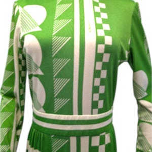May include: A green and white patterned dress with long sleeves. The dress features a collared neckline and a fitted waist. The pattern includes geometric shapes such as triangles, squares, and circles. The dress is a vintage style.