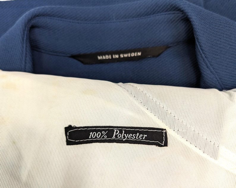 Puede incluir: Primer plano de una etiqueta de tela blanca con texto negro que dice "100% Poli&eacute;ster". La etiqueta est&aacute; unida a una tela azul con una etiqueta negra que dice "Hecho en Suecia".