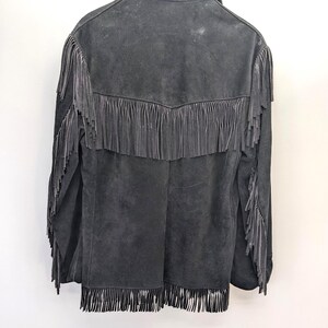May include: A dark grey suede jacket with fringe detailing. The jacket has fringe across the back yoke, sleeves, and hem. It is hanging on a wooden hanger against a white background.
