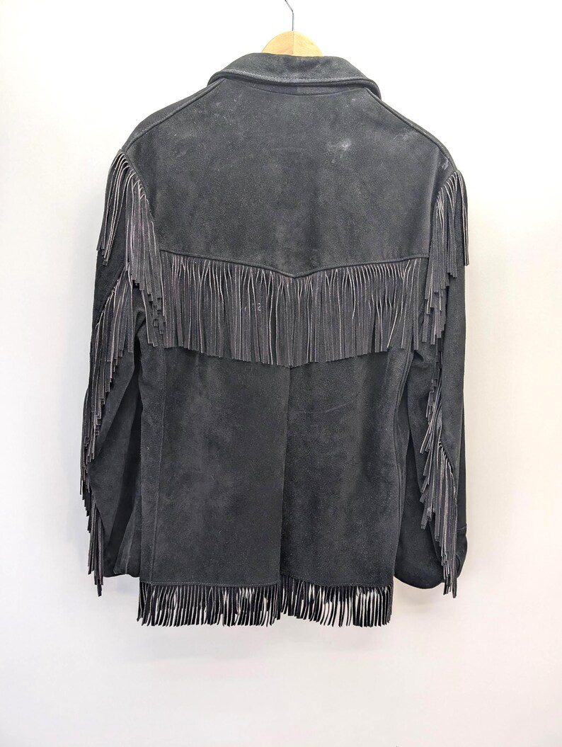 May include: A dark grey suede jacket with fringe detailing. The jacket has fringe across the back yoke, sleeves, and hem. It is hanging on a wooden hanger against a white background.