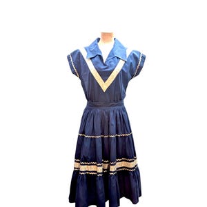 Puede incluir: Un vestido azul marino con cuello blanco y ribetes blancos. El vestido tiene una falda de capas con ribetes de encaje blanco.