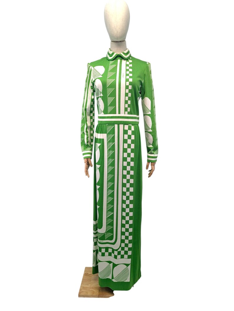 May include: A long, green dress with a white geometric pattern. The dress features long sleeves, a collared neckline, and a fitted waist. The pattern includes squares, circles, and lines. The dress is displayed on a mannequin.