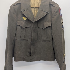 May include: An olive drab military jacket featuring a collared neckline, two front chest pockets with flaps, and a front zipper closure. The left sleeve displays a patch with the number 15 and chevron insignia, while a small yellow pin is on the chest.
