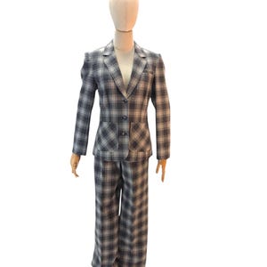 May include: A gray and white plaid suit, consisting of a blazer and matching trousers, displayed on a wooden mannequin. The blazer has a two-button closure and two front pockets. The trousers are wide-legged.