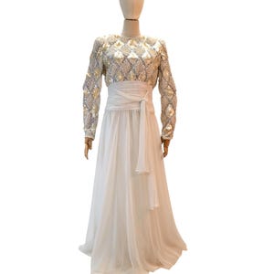 May include: A long, white formal gown on a mannequin. The bodice features a pattern of diamond-shaped embellishments in silver and gold. The long sleeves are also embellished. A white sash cinches the waist, and the skirt is a flowing, sheer fabric.