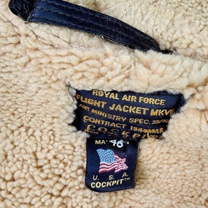 Może przedstawiać: Czarna sk&oacute;rzana metka z białym tekstem, kt&oacute;ry brzmi "ROYAL AIR FORCE FLIGHT JACKET MKVI AIR MINISTRY SPEC. 39/76 CONTRACT 19494BAR MA 46" z małą amerykańską flagą i tekstem "U.S.A. COCKPITTM".