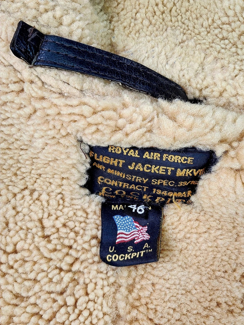Może przedstawiać: Czarna sk&oacute;rzana metka z białym tekstem, kt&oacute;ry brzmi "ROYAL AIR FORCE FLIGHT JACKET MKVI AIR MINISTRY SPEC. 39/76 CONTRACT 19494BAR MA 46" z małą amerykańską flagą i tekstem "U.S.A. COCKPITTM".