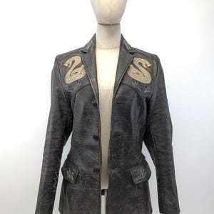 May include: A black leather jacket with a button-up front and two snake embroidered designs on the chest. The jacket has a distressed look and features decorative stitching on the pockets and sleeves.