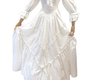May include: A white off-the-shoulder dress with long sleeves and a full skirt. The dress features a bow at the neckline, ruffled details, and a tiered skirt. The sleeves have ruffled cuffs. The dress is displayed on a mannequin.